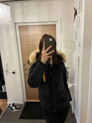 Svart Parajumpers parkas med pälskrage - Säljer en svart parkas från Parajumpers med stor fluffig pälskrage i beige och svart. Jackan har dragkedja (OBS dragkedjan har gått sönder lite men fungerar fortfarande om man inte är för hård hänt), stora fickor och den klassiska gula remmen vid kragen. Perfekt för kalla dagar och riktigt snygg streetstil.