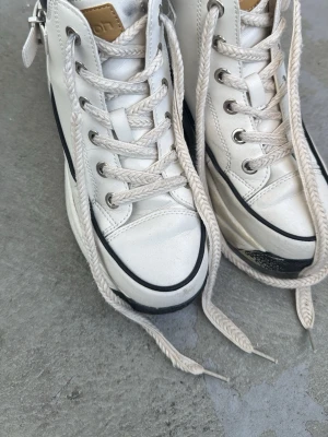 Vita chunky high top sneakers - Säljer ett par vita chunky high top sneakers med svart detalj vid hälen och silverfärgat spänne på sidan. Skorna har tjock sula, vita snören och svarta linjer längs sulan. Perfekta för dig som gillar streetstyle och vill sticka ut.
