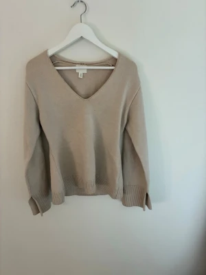 Beige stickad tröja med v-ringning - En stilren beige stickad tröja från H&M med djup v-ringning och ribbade muddar vid ärmslut och nederkant. Tröjan har en avslappnad passform och är perfekt för lager-på-lager. Tillverkad i mjukt material som känns skönt mot huden.
