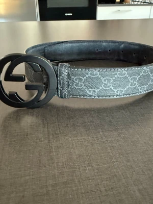 Gucci bälte - Snyggt Gucci bälte i grått med klassiskt GG-mönster och stort svart GG-spänne. Bältet har en bred design och är tillverkat i äkta läder med textil på utsidan. Perfekt accessoar för att lyfta din outfit.