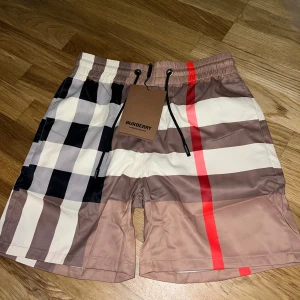 Rutig Burberry shorts  - Snygga Burberry shorts i klassiskt rutigt mönster med beige, brun, svart, vit och röd färg. De har elastisk midja med svart snörning och meshfoder på insidan. Perfekt för sommaren och har en loose passform som ger skön vibe. Storlek M men passar S
