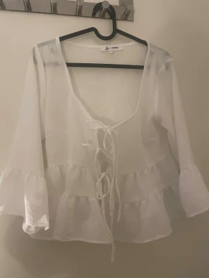 Vit volangblus med knytning - Snygg vit blus från Sisters Point med volangdetaljer och knytning framtill. Blusen har vida ärmar och djup urringning, perfekt för en trendig och luftig look. Tillverkad i ett lätt och transparent material.