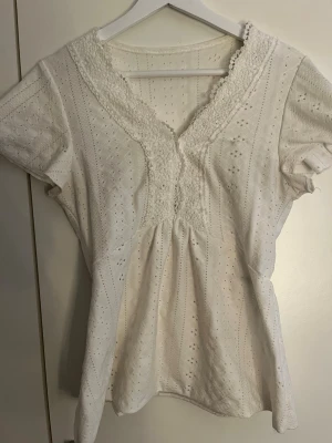 Vit blus med spets och v-ringning - Söt vit blus med korta ärmar, v-ringad hals och spetsdetaljer längs ringningen. Blusen har broderade hålmönster och resår i midjan för en feminin siluett. Perfekt till jeans eller kjol för en fräsch look.