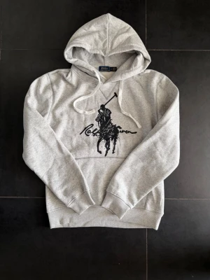 Grå hoodie från Polo Ralph Lauren - Snygg grå hoodie från Ralph Lauren med stor svart polospelare och logga på bröstet. Klassisk känguruficka framtill, ribbade muddar och justerbar huva med vita snören. Tillverkad i mjukt sweatshirtmaterial som är perfekt för chill dagar. Storlek M men sitter mer åt S hållet ✅