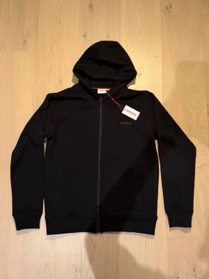 Svart hoodie med dragkedja från Hugo - Svart hoodie från Hugo med dragkedja framtill och diskret logotyp på bröstet. Modellen har huva, ribbade muddar och två sidofickor. Materialet är mjukt och bekvämt, perfekt för en avslappnad look. Passar dig som gillar stilrena basics.