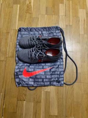 Grå Nike fotbollsskor med väska - Snygga grå fotbollsskor från Nike med röda detaljer och klassisk Nike-logga. Skorna har plastdobbar och är tillverkade i syntetmaterial. Medföljer en grå Nike-väska med tryck och röd logga. Perfekta för spel på konstgräsplan. Har bara använt dem en gång och de var för små så jag tänker sälja dem jag köpte dem 2999.
