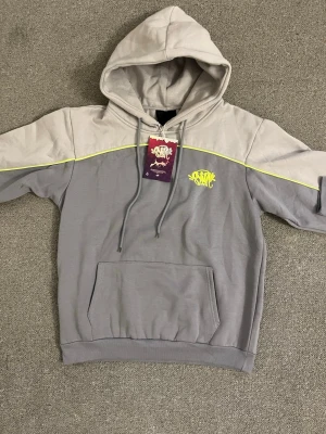 Grå hoodie från Syna worldmed neon-detaljer - Snygg tvåfärgad hoodie från SXTN i ljusgrått och mörkgrått med neon-gul logga på bröstet och matchande neon-gul linje över bröstet. Klassisk känguruficka framtill och justerbar huva med dragsko. Perfekt för en streetwear-look.