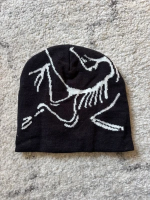 Svart mössa med vitt skelettmönster - Cool svart mössa med ett vitt grafiskt skelettmönster på framsidan. Mössan är stickad och har en klassisk, något slouchy passform. Perfekt för dig som vill sticka ut med en unik accessoar till hösten eller vintern.