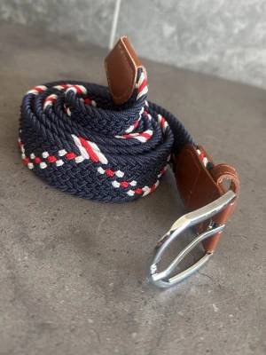 Randigt flätat bälte med läderdetaljer - Snyggt flätat bälte i marinblått med röda och vita ränder. Dess skick är mycket bra 8/10, vet inte exakta längden men kom dm om ni vill ha den exakta längden! Kom även dm om ni har några frågor eller om ni vill ha mer bilder!