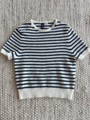 Randig stickad tröja från H&M - Säljer en snygg stickad tröja från H&M med horisontella ränder. Tröjan har korta ärmar, rund halsringning och ribbade muddar. Materialet är mjukt och ser ut att vara ullblandning. Perfekt för en stilren och avslappnad look.