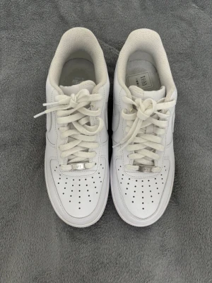 Nike Air Force 1 vita sneakers - Klassiska helvita Nike Air Force 1 sneakers i lågt utförande. Skorna har ovandel i slätt läder, perforerade detaljer på tån och vit sula. Ikonisk Nike Swoosh på sidan och metallbricka med AF1 på snörningen. Perfekta för en clean och stilren look.