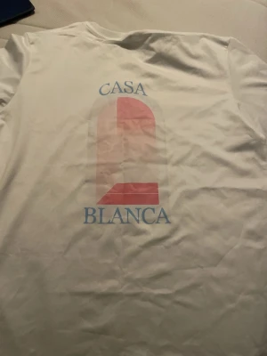 Vit t-shirt från Casablanca - Vit t-shirt med tryck på ryggen från Casablanca. Trycket består av en rosa grafisk båge och texten 'CASA BLANCA' i ljusblått. Klassisk passform och rund hals. Perfekt för en stilren och trendig look.