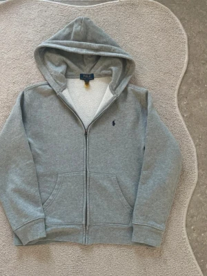 Grå hoodie med dragkedja från Polo Ralph Lauren - Snygg grå hoodie från Polo Ralph Lauren med dragkedja framtill och klassisk huva. Storlek barn 14-16 år.