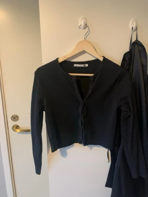 Svart cardigan från lager  157 - Klassisk svart cardigan från 759 i storlek L. Tröjan har rund halsringning, knäppning framtill med svarta knappar och långa ärmar. Perfekt att slänga över en t-shirt eller linne för en enkel och stilren look.