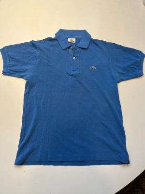 Blå pikétröja från Lacoste - Klassisk blå pikétröja från Lacoste med broderad krokodillogga på bröstet. Tröjan har korta ärmar, krage och knäppning med två knappar framtill. Perfekt för en stilren och sportig look.