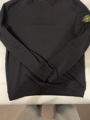 Svart sweatshirt från Stone Island - Säljer en svart sweatshirt från Stone Island med den klassiska logotypen på vänster ärm. Tröjan har rund halsringning, långa ärmar och ribbade muddar. Perfekt för dig som gillar streetwear och vill ha en stilren look.