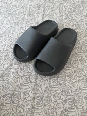 YEEZY Adidas - Säljer ett par gråa slides från YEEZY med stilren och chunky design. Tofflorna har öppen tå och är gjorda i ett mjukt och flexibelt syntetmaterial. Perfekta att glida i hemma eller på stranden. Adidas-logga syns diskret på sulan. OBS tofflorna säljs utan låda! 