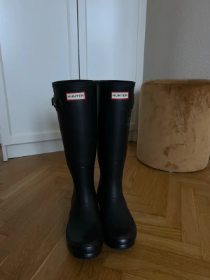 Svarta höga gummistövlar Hunter - Klassiska svarta höga gummistövlar från Hunter med justerbart spänne på sidan och logotyp framtill. Tillverkade i slitstarkt gummi och perfekta för regniga dagar. Snygg och tidlös design som funkar till många olika stilar. Väldigt bra skick. Säljs pga passar inte.