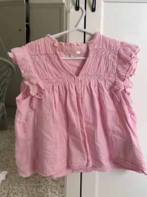 Rosa volangblus från Gina Tricot - Söt ljusrosa blus från Gina Tricot med volangdetaljer vid ärmöppningarna och smock upptill. Blusen har v-ringning och är tillverkad i ett mjukt, tunt bomullstyg. Perfekt för en somrig och avslappnad look.