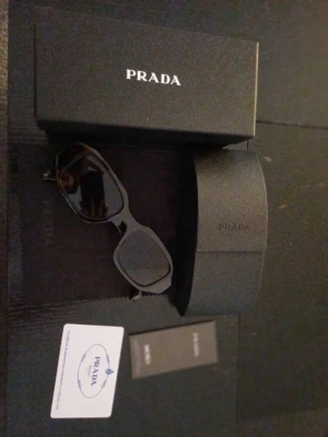 Svarta solglasögon från Prada - Snygga svarta solglasögon från Prada med rektangulära bågar och mörka glas. Kommer med originalfodral, låda och äkthetskort. Perfekt accessoar för dig som vill ha en stilren och exklusiv look.