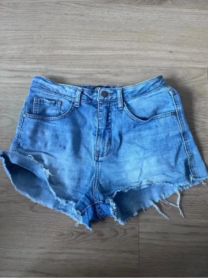 Blå jeansshorts med fransig kant - Säljer ett par klassiska blå jeansshorts från Wrangler med låd midja och rå, fransig kant. Shortsen har fem fickor och knappgylf. Perfekta för varma dagar och ger en avslappnad vibe.
