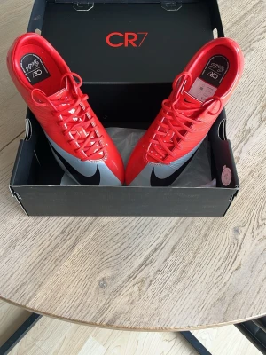 Nike CR7 Mercurial fotbollsskor röd/silver - Dessa nike CR7 Mercurial fotbollsskor i rött och silver med svarta detaljer. Skorna har en smal, aerodynamisk form och är tillverkade i syntetmaterial för bästa bollkontroll. Och gjordes bara ett limiterat antal. Utrustade med fasta dobbar för spel på gräs. Perfekta för dig som vill sticka ut på planen. Priset är inte hugget i sten