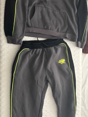 Grått loungewear-set med neonränder - Mysigt tvådelat loungewear-set i mörkgrått med svarta detaljer och neonfärgade ränder längs ärmar och ben. Setet består av en sweatshirt med magficka och matchande byxor med dragsko i midjan. Perfekt för chill dagar hemma.