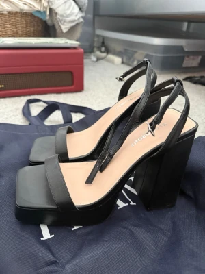 Svarta chunky pumps från Glamorous - Snygga svarta pumps från Glamorous med chunky klack och fyrkantig tå. Skorna har en stilren rem över tårna och justerbar ankelrem med silverfärgat spänne. Tillverkade i konstläder med beige innersula för extra komfort.