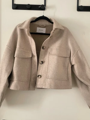 Beige kappa från Stradivarius  - Stilren beige kappa  i ullmix från Stradivarius. Modellen är boxig och croppad med stora bröstfickor och knappar framtill. Perfekt att slänga över en hoodie eller t-shirt för en chill look. Klassisk krage och långa ärmar med knapp vid ärmslut.