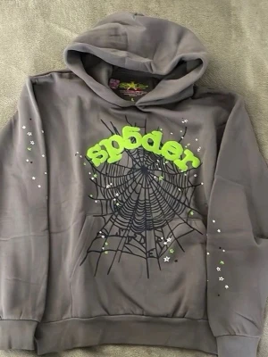 Grå hoodie med spindelnätsmönster - Snygg grå hoodie med huva och ett stort svart spindelnätsmönster på bröstet. Texten 'sp6der' i neon-grönt ger en cool kontrast. Små vita och gröna stjärnor pryder ärmarna för extra detalj. Perfekt för dig som gillar streetwear och unika prints.