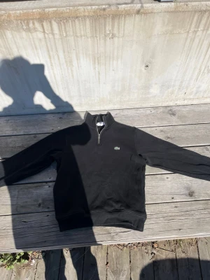 Svart halfzip hoodie från Lacoste - Säljer en svart hoodie från Lacoste med klassisk halv dragkedja och det ikoniska gröna krokodilmärket på bröstet. Hoodien har ribbade muddar och är tillverkad i mjukt bomullsmaterial. Perfekt för en clean och stilren look.