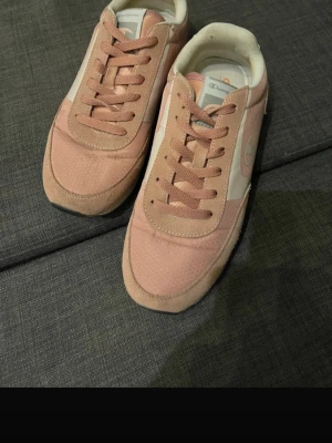Rosa och vita sneakers från Champion - Säljer ett par snygga sneakers från Champion i ljusrosa och vitt. Skorna har klassisk snörning, rund tå och är tillverkade i en mix av mocka och syntetmaterial. Perfekta för dig som vill ha en sportig och trendig look.