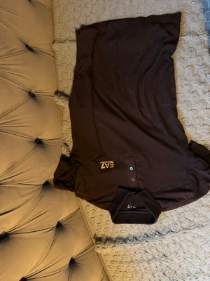 Svart pikétröja från EA7 Emporio Armani - Snygg svart pikétröja från EA7 Emporio Armani med klassisk krage och tre knappar framtill. Tröjan har guldfärgad logga på bröstet och diskreta guldstreck på ryggen. Tillverkad i mjukt bomullsmaterial som känns skönt mot huden.    Endast använd fåtal gånger.