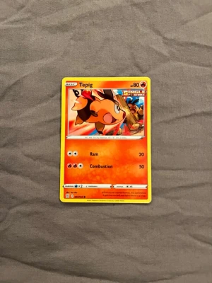 Pokémonkort Tepig Single Strike - Säljer ett Pokémonkort med Tepig från Single Strike-serien. Kortet har en färgstark design med Tepig i orange, gult och svart mot en röd bakgrund. Det är på engelska och visar attackerna Ram och Combustion. Perfekt för samlare eller Pokémonfans.