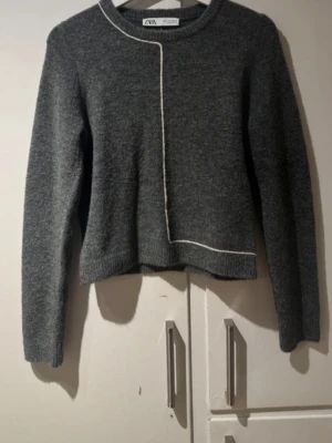 Grå stickad tröja från Zara - Mysig grå stickad tröja från Zara med lång ärm och rund hals. Tröjan har en vit dekorativ linje som går från halsen och ner över framsidan, vilket ger en cool och modern look. Perfekt att matcha med jeans eller kjol.