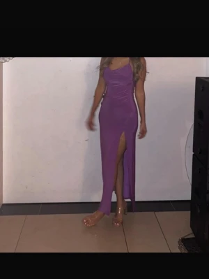 Lila lång klänning med slits - Snygg lila långklänning med en axel och hög slits framtill. Klänningen har figurnära passform och är tillverkad i ett glansigt material som ger en lyxig känsla. Perfekt för dig som vill sticka ut med en trendig och elegant look.