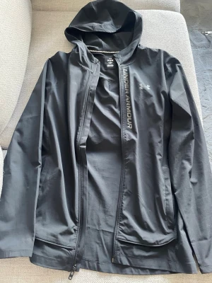 Svart vindjacka från Under Armour - Snygg svart vindjacka från Under Armour med huva och dragkedja framtill. Jackan har logga och text på bröstet samt två sidofickor. Perfekt för blåsiga dagar och sportiga outfits.