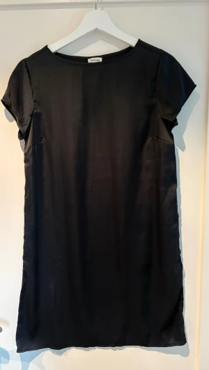Svart kortärmad klänning från Bubbleroom - En stilren svart klänning från Bubbleroom med en känsla av satin. Klänningen har korta ärmar, rund halsringning och rak passform. Perfekt för dig som gillar minimalistisk och tidlös stil. Axelmått 35 cm och längd 89 cm. Aldrig använd.