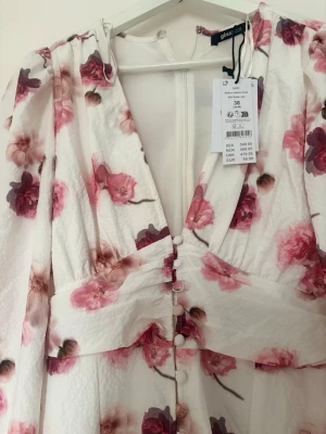 Vit blommig klänning från Gina Tricot - Romantisk vit klänning från Gina Tricot med rosa och vinröda blommor. Klänningen har v-ringning, knappar framtill och långa ärmar. Tillverkad i ett lätt, strukturerat material som ger en luftig känsla. Perfekt för vår och sommar!