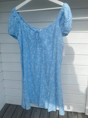 Blå blommig kortklänning med puffärm - Söt ljusblå kortklänning med små vita blommor, puffärmar och knytning fram i halsen. Klänningen har en luftig passform och är perfekt för varma dagar. Tillverkad i ett lätt bomullstyg som känns skönt mot huden.