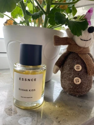 ESSNCE Sugar Kiss Eau de Parfum - ESSNCE Sugar Kiss Eau de Parfum i en stilren, rund glasflaska med svart lock. Parfymen har en ljusgul vätska och etiketten är enkel och modern. Perfekt för dig som gillar söta dofter och vill ha en snygg parfymflaska på hyllan.