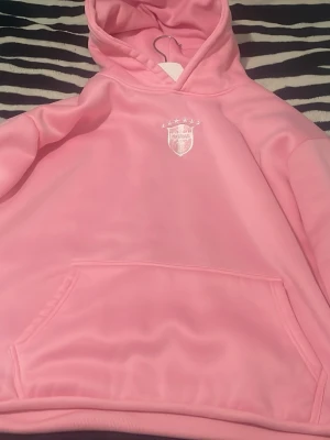 Rosa hoodie med broderad emblem - Snygg rosa hoodie med broderad emblem på bröstet och fem stjärnor ovanför. Hoodien har en stor känguruficka framtill och en rejäl huva. Materialet är mjukt och känns skönt mot huden, perfekt för att chilla eller hänga med kompisar.