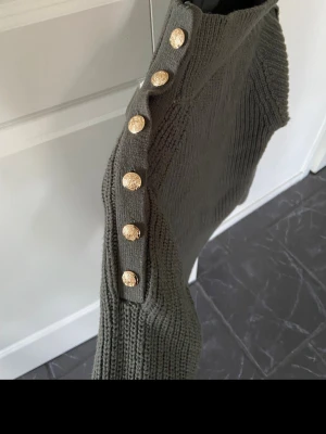 Olivgrön stickad tröja med guldknappar - Snygg olivgrön stickad tröja med ribbad struktur och dekorativa guldfärgade knappar längs axlarna. Tröjan har hög hals och långa ärmar, perfekt för att hålla värmen. Passar dig som gillar trendiga detaljer och en klassisk look.