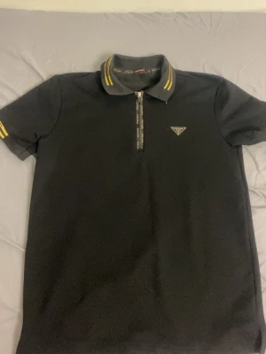 Svart pikétröja med dragkedja från Prada - Cool svart pikétröja från Prada med dragkedja framtill och klassisk krage. Tröjan har gula detaljer på kragen och ärmarna samt den ikoniska metall-loggan på bröstet. Perfekt för dig som vill ha en stilren och exklusiv look.