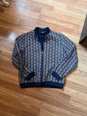 Blå mönstrad ziptröja från Lacoste - Snygg blå ziptröja från Lacoste med vitt geometriskt mönster och klassisk krokodillogga på bröstet. Tröjan har ribbade muddar, hög krage och dragkedja framtill. Perfekt för en sportig och trendig look. Inköpt på rea på Kids Brand Store för 1394 kr. Se kvitto. 