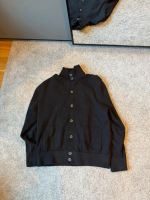 Svart kofta  - Stilren svart kofta från Jules med hög krage och stora knappar framtill. Två praktiska fickor framtill och ribbade muddar vid ärmslut och nederkant. Perfekt för lager-på-lager och enkel att matcha med andra plagg. knappt änvänd