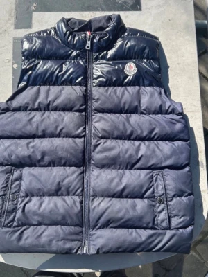 Mörkblå dunväst från Moncler - Snygg mörkblå dunväst från Moncler med glansig ovandel och klassisk logga på bröstet. Västen har ståkrage, dragkedja framtill och två fickor med knappstängning. Perfekt för lager-på-lager och streetstyle. Priset kan diskuteras. Storleken är 158 men passar Sture.