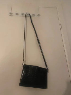 Svart axelväska i croco-mönster - Snygg svart axelväska med croco-mönstrad yta och justerbar axelrem. Väskan har en stilren, fyrkantig form och stängs med dragkedja. Perfekt för dig som gillar trendiga accessoarer med lite edge.