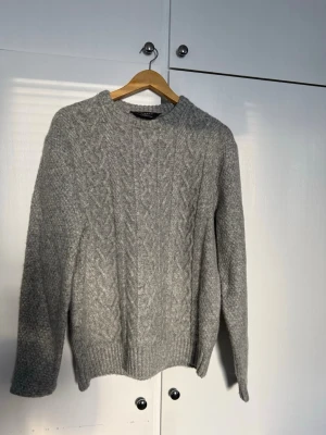 Grå kabelstickad tröja från PRIMARK - Säljer en grå kabelstickad tröja från Lexington med rund halsringning och ribbade muddar. Tröjan har ett klassiskt flätmönster framtill och är gjord i ett mjukt, värmande material. Perfekt för kyliga dagar och enkel att matcha med jeans eller kjol.
