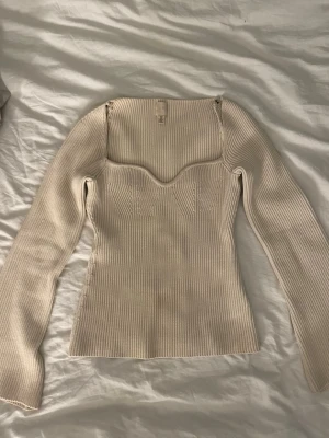 Ribbad beige långärmad topp - Trendig ribbad topp i beige med lång ärm och markerad byst. Fyrkantig urringning ger en modern look och den åtsittande passformen framhäver figuren. Perfekt att styla med jeans eller kjol för en clean och snygg outfit.